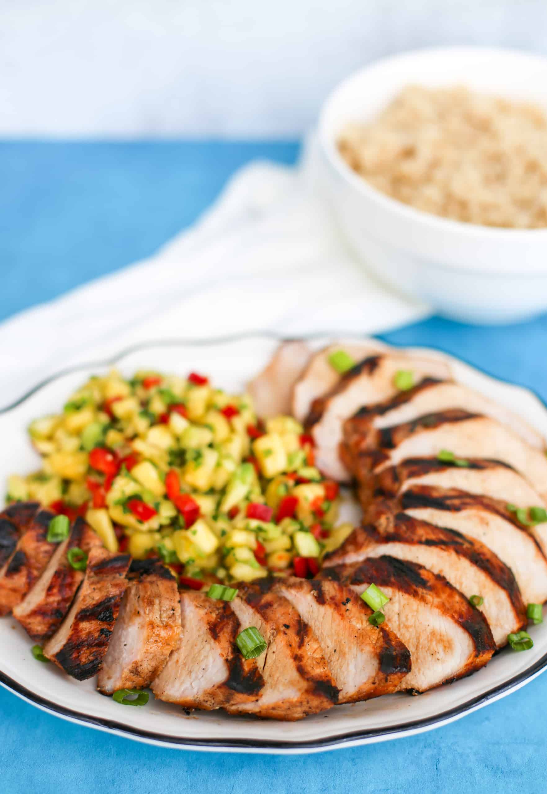 Cooked and sliced grilled pork tenderloin on a platter with Pineapple Salsa on the side and brown rice in a bowl next to it.