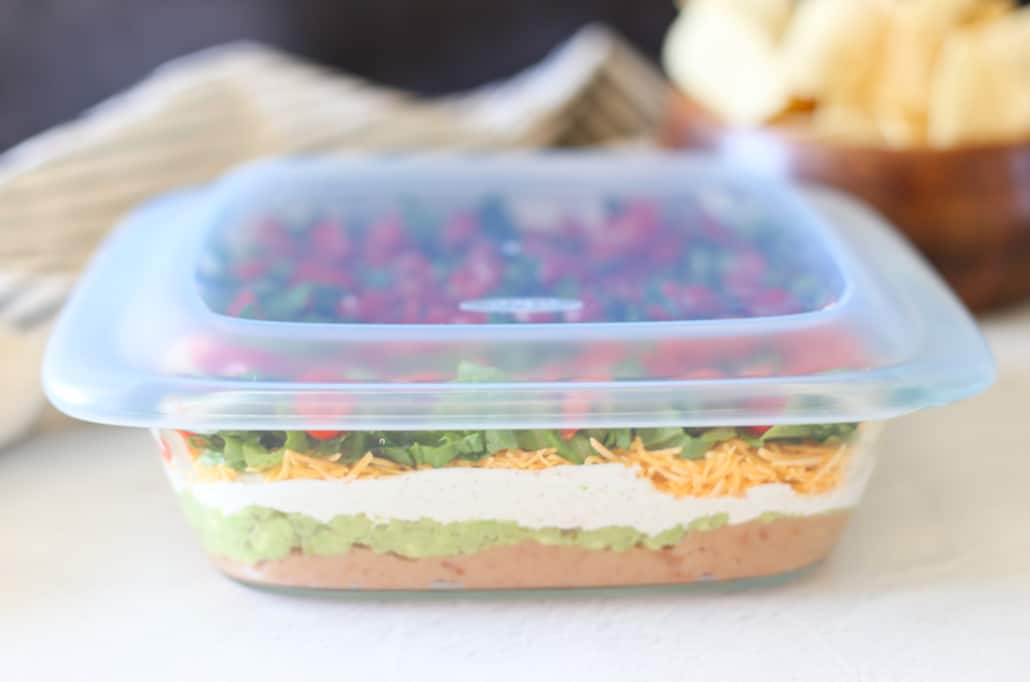 Layered taco dip in a glass dish with a lid