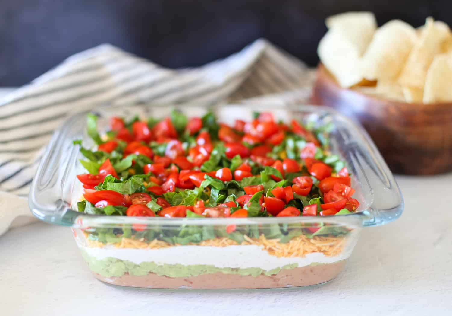 Layered taco dip in a glass 8x8 pan with refried beans on the bottom and chopped tomatoes and lettuce on top.