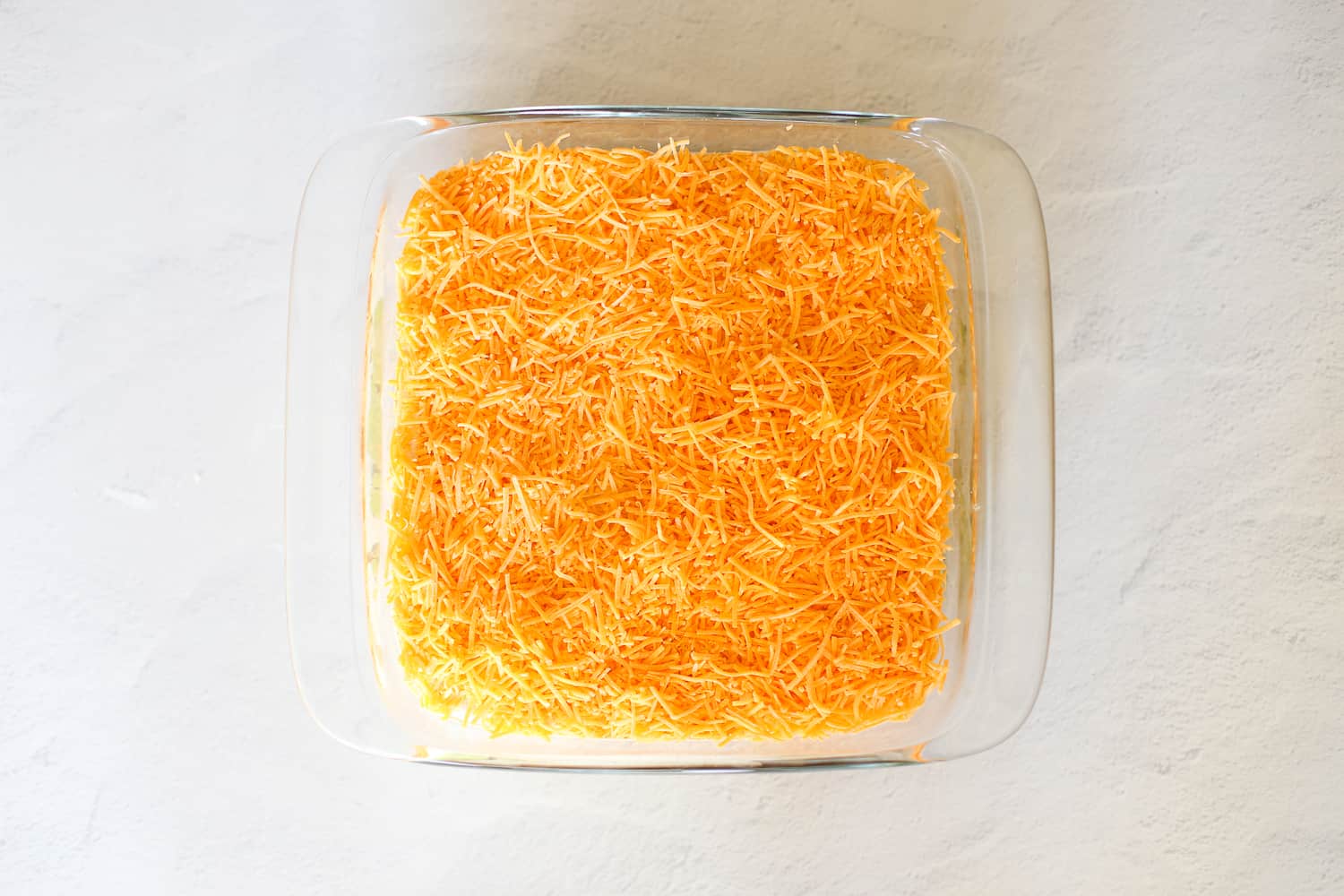 Shredded cheese covering layers of refried beans, guacamole and sour cream in a baking dish.