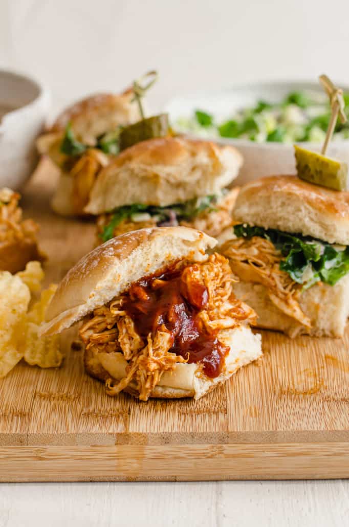 Bbq chicken sliders on a serving board with slaw and chips on the side. 