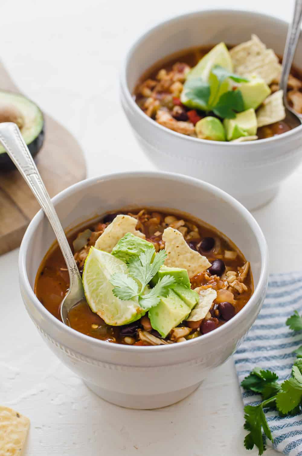 Two white bowls with chicken taco soup and toppings set on a table with spoons.