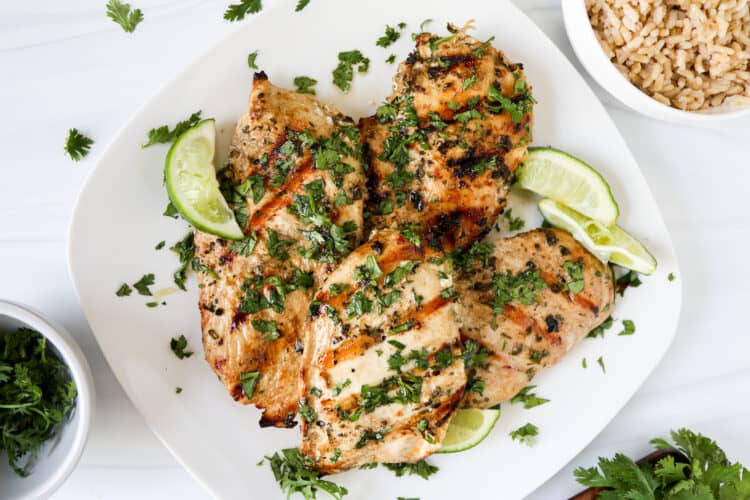 Grilled cilantro lime chicken on a square serving dish with lime wedges and chopped cilantro.