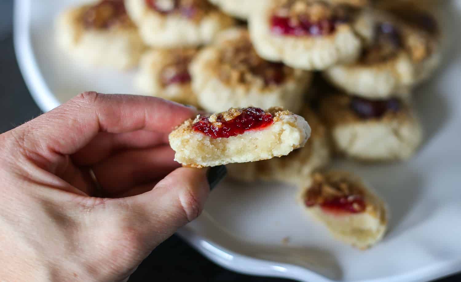 Hand holding a raspberry crumble cookie with a bite taken out of it and a platter of cookies in the background.