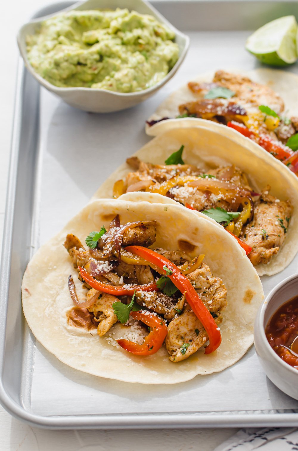 vertical shot of chicken fajitas lined up on a tray with salsa and guacamole in small bowls on the side