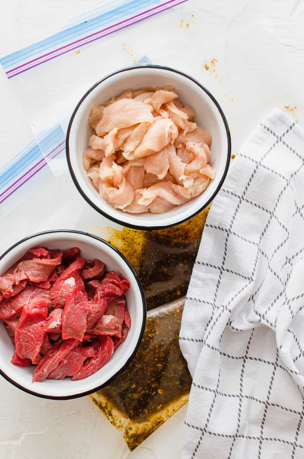 process shot of thinly sliced steak and thinly sliced chicken in separate bowls along with two freezer bags of the fajita marinade