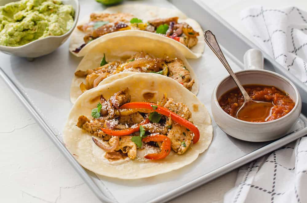 chicken fajitas lined up on a tray with salsa and guacamole in small bowls on the side