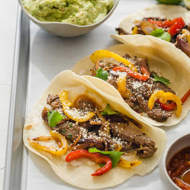 vertical shot of steak fajitas lined up on a tray with guacamole and lime wedges in the background