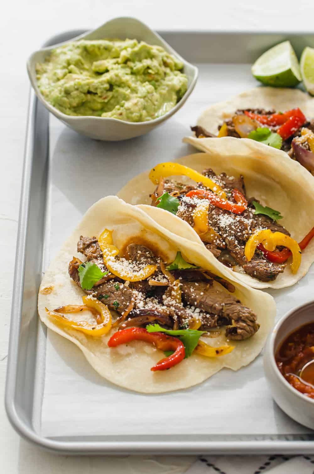 vertical shot of steak fajitas lined up on a tray with guacamole and lime wedges in the background