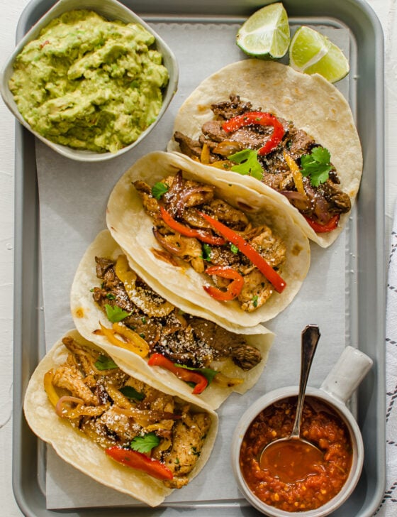 vertical shot of steak and chicken fajitas lined up on a tray with salsa, lime, and guacamole on the side