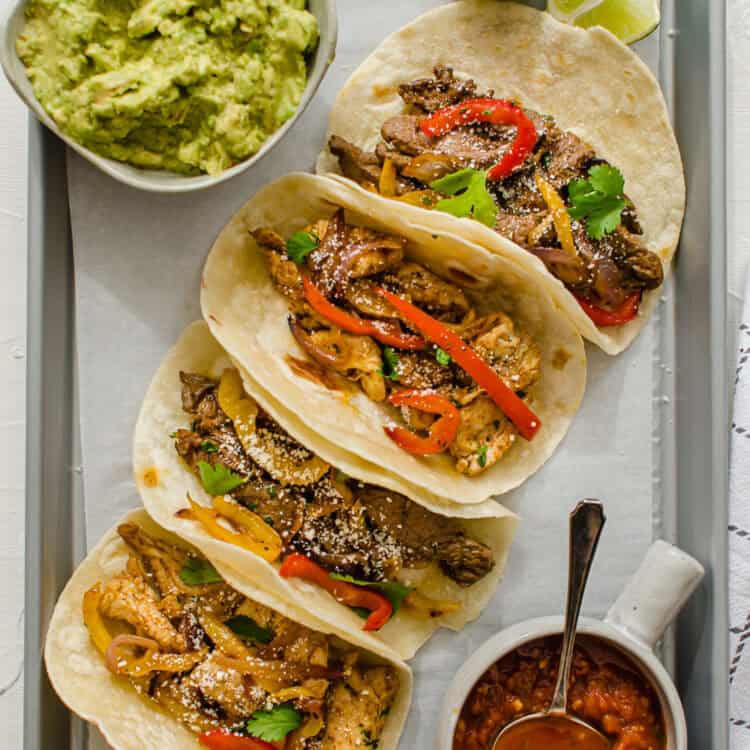 Steak and chicken fajitas lined up on a tray with salsa, lime, and guacamole on the side.