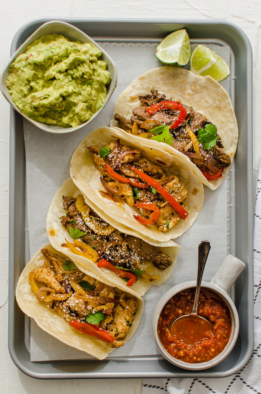 vertical shot of steak and chicken fajitas lined up on a tray with salsa, lime, and guacamole on the side