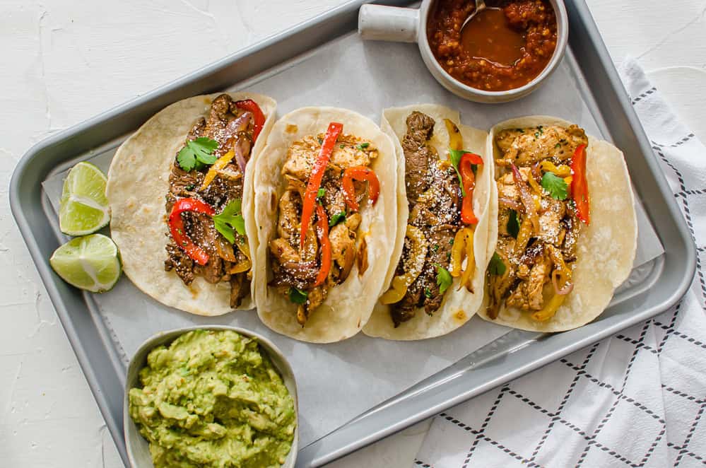 Steak and chicken fajitas lined up on a tray with salsa, lime, and guacamole on the side.