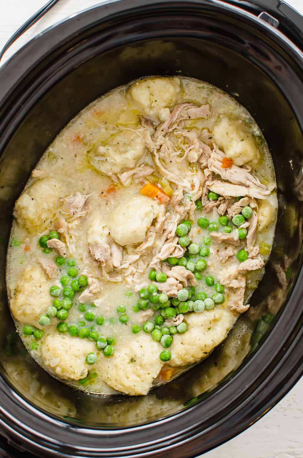 Frozen peas and shredded chicken being stirred in a slow cooker for chicken and dumplings.