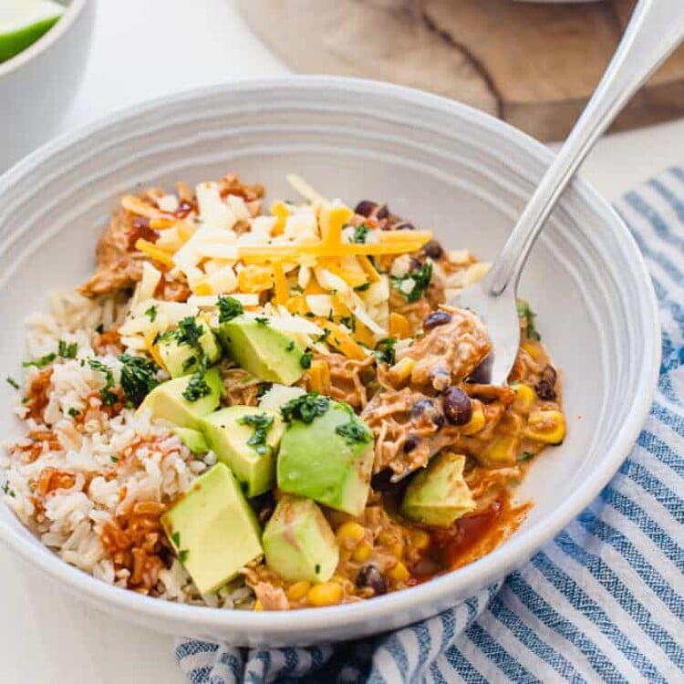 Crockpot Mexican Chicken served in a white bowl with avocado chunks, shredded cheese, and chopped cilantro on top.