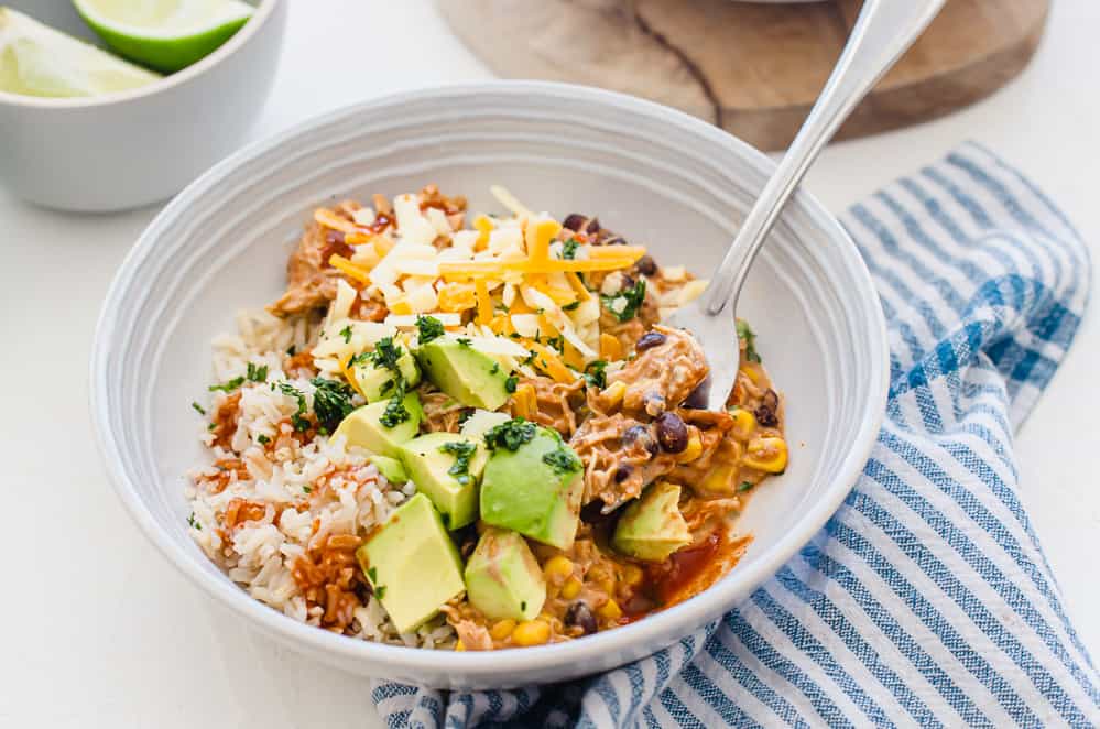 Crockpot Mexican Chicken served in a white bowl with avocado chunks, shredded cheese, and chopped cilantro on top.