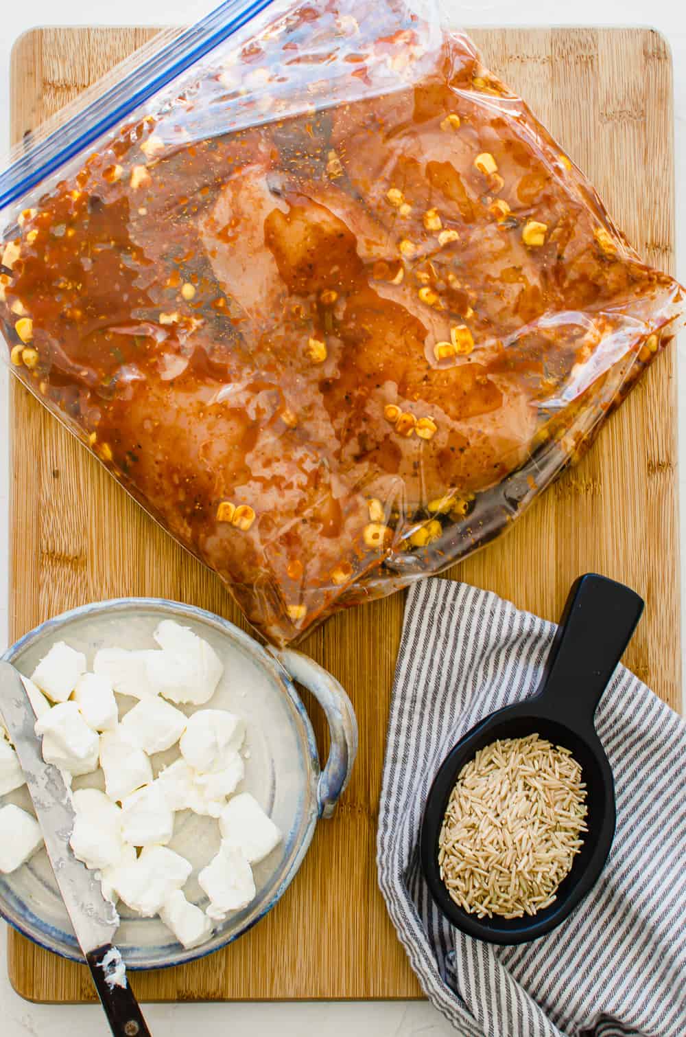 Mexican chicken ingredients in a freezer bag lying flat on a wooden cutting board with cubed cream cheese and uncooked brown rice next to it.