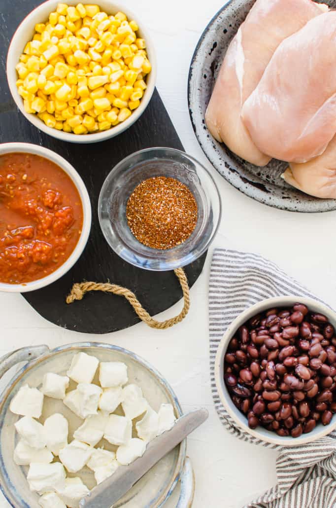 ingredients for mexican chicken in separate bowls including chicken breasts, corn, salsa, taco seasoning, black beans, and cream cheese