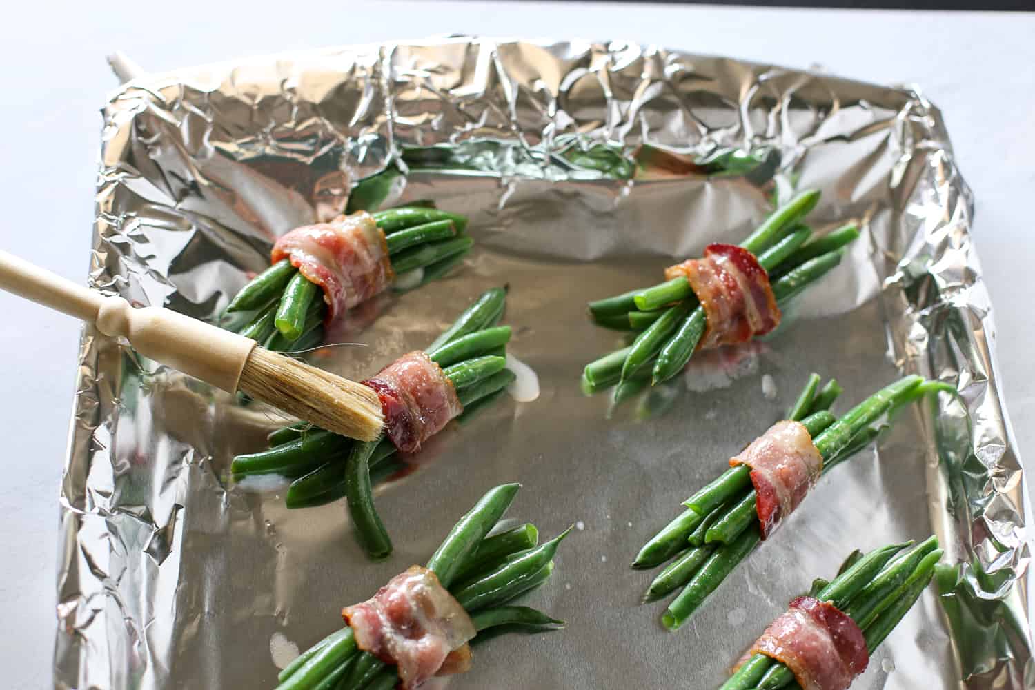 Green beans wrapped in bundles with bacon sitting on a foil-lined baking sheet being brushed with garlic butter.