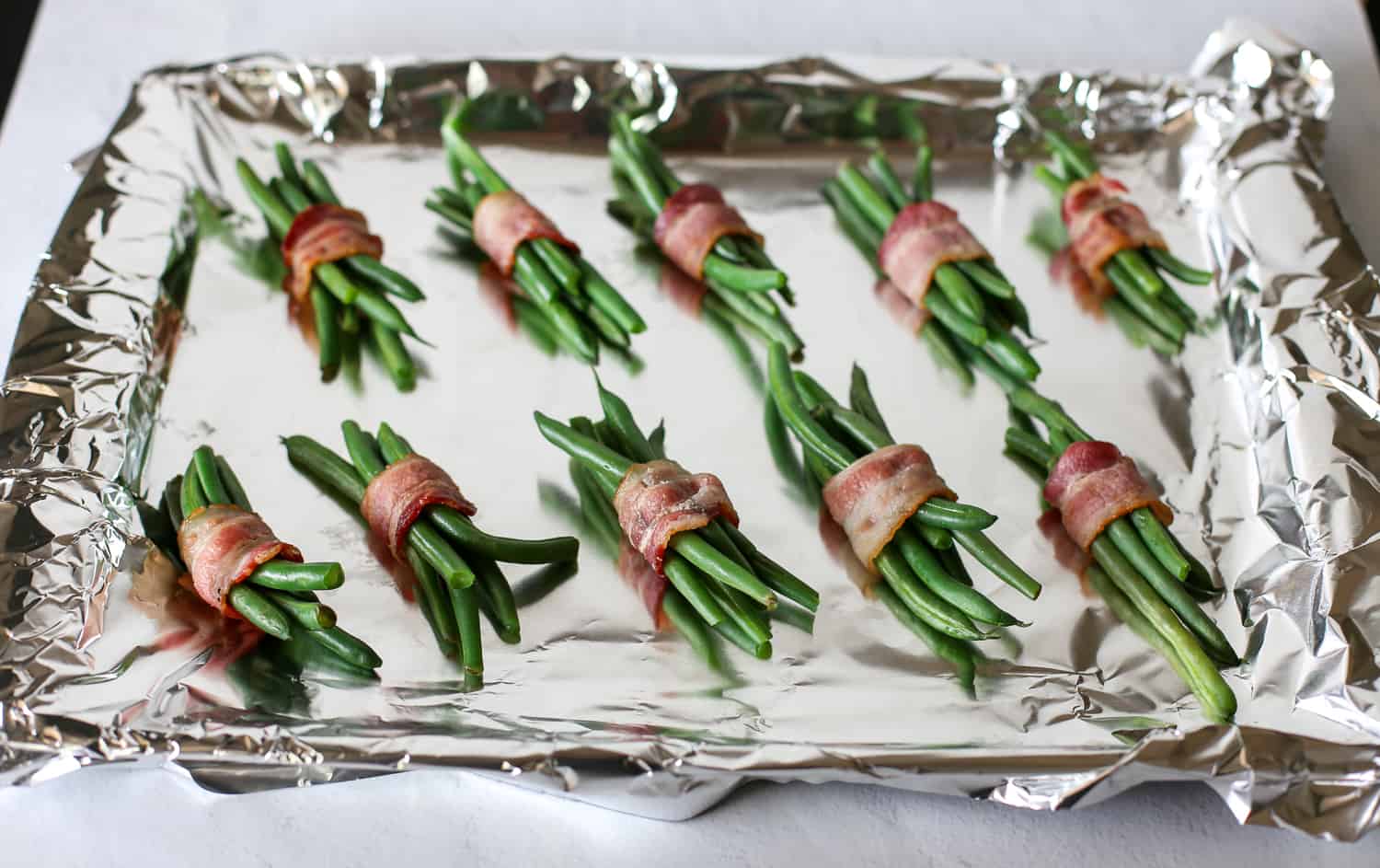 Fresh, trimmed green beans wrapped in bacon sitting on a foil-covered baking sheet.