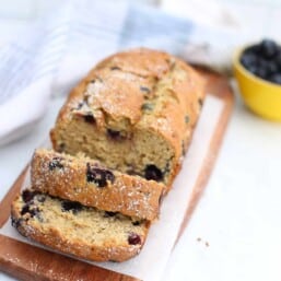 Blueberry banana bread on a wooden cutting board with some of it sliced.