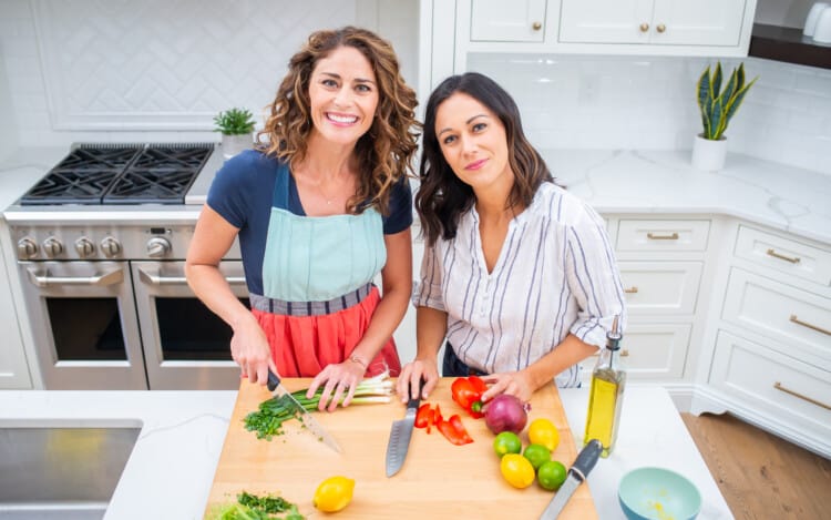 Rachel Tiemeyer and Polly Conner in a kitchen chopping vegetables on a wooden cutting board.