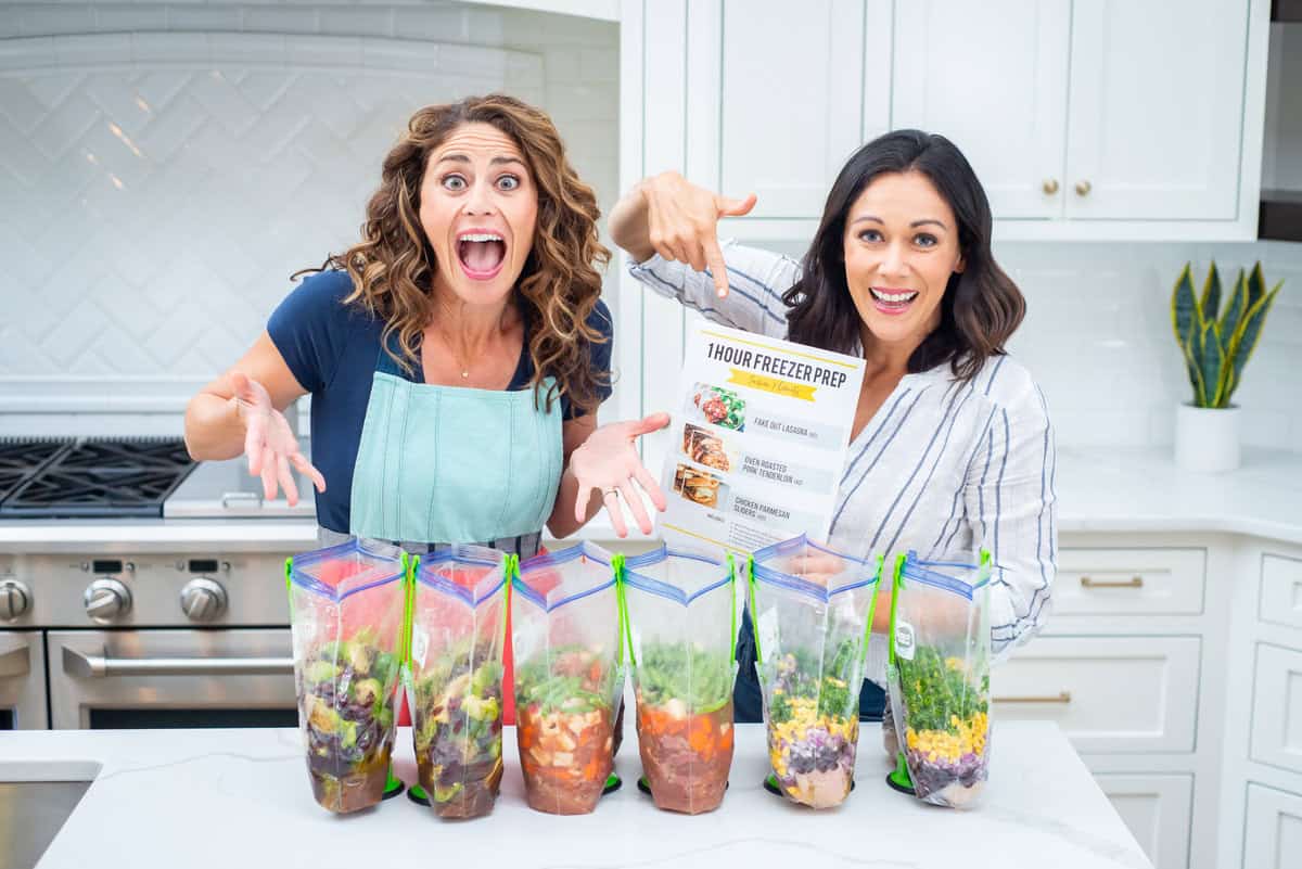 Rachel and Polly from Thriving Home holding a copy of their 1 Hour Freezer Prep session standing in front of six gallon-size bags with different freezer meals in them.