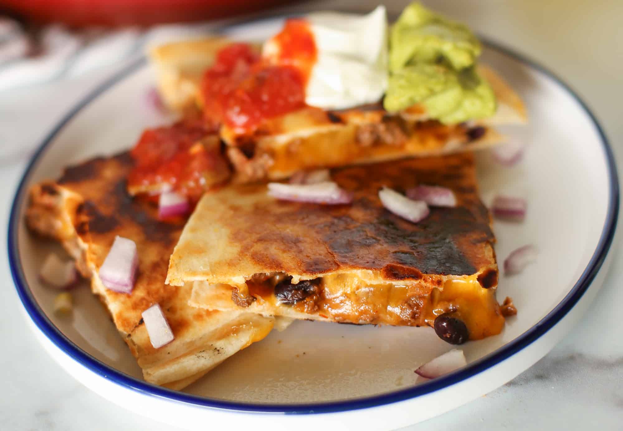 Beef quesadilla cut up with salsa, sour cream, guacamole, and chopped onion on top. 