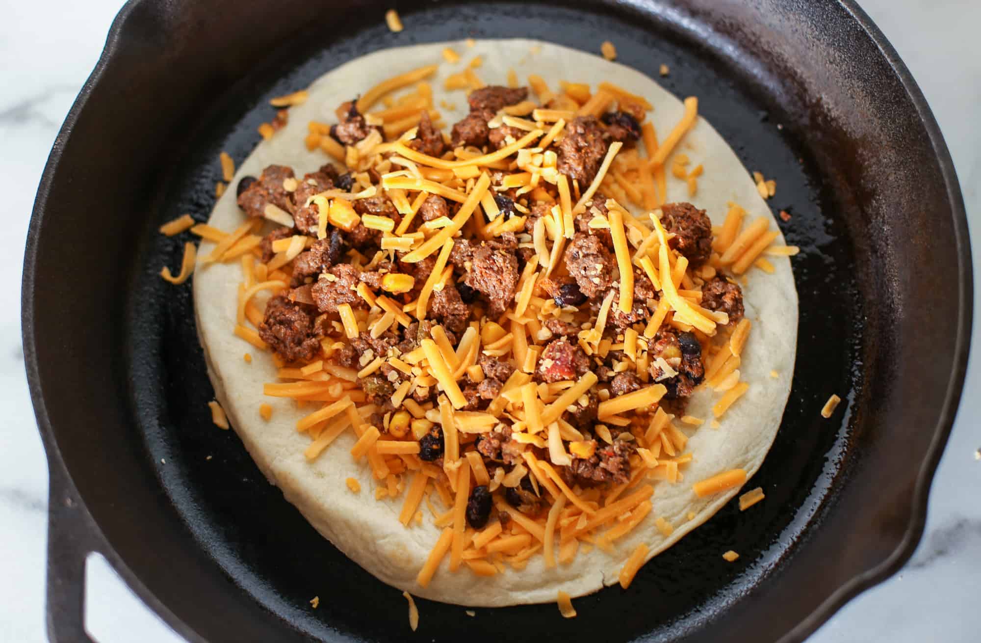 Tortilla with ground beef mixture and shredded cheese in a cast iron skillet.