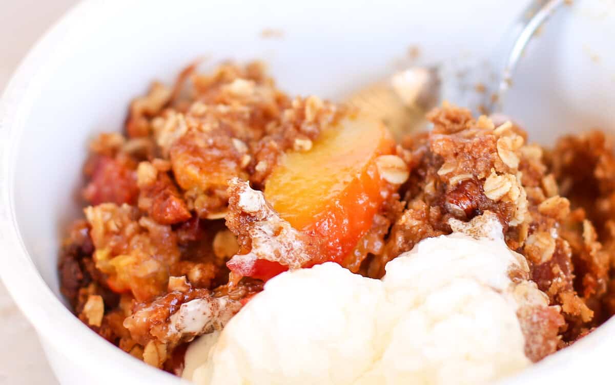 Peach crisp served in a white bowl with vanilla ice cream.