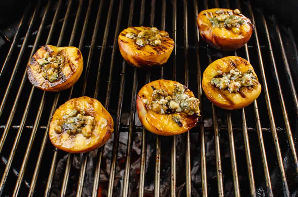 Grilled peach halves on a grill with Gorgonzola cheese, balsamic drizzle, and honey on top of each one.