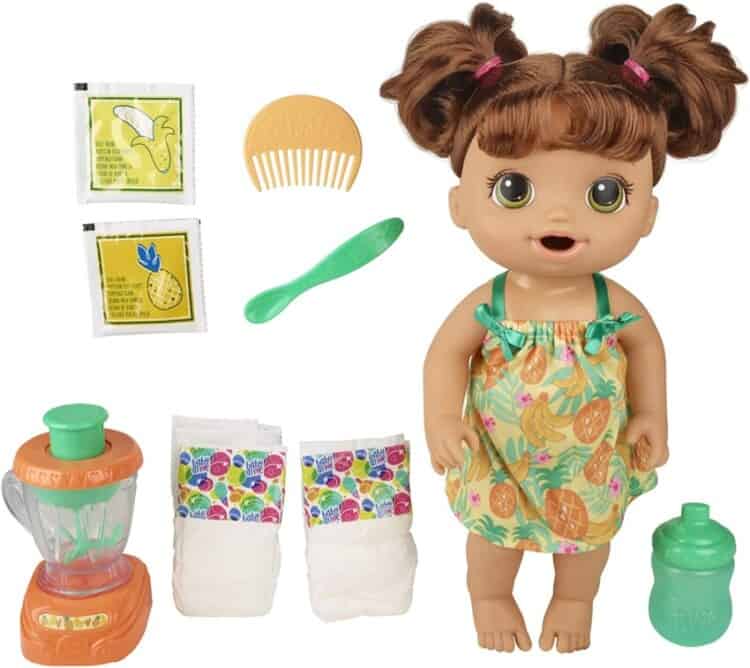 A brown-haired Baby Alive doll with accessories like diapers, a comb, a bottle and other items sitting around the doll.
