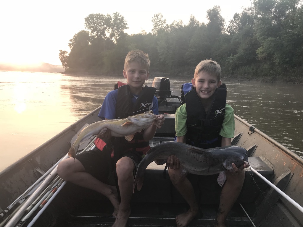 Boys holding large catfish in a boat.