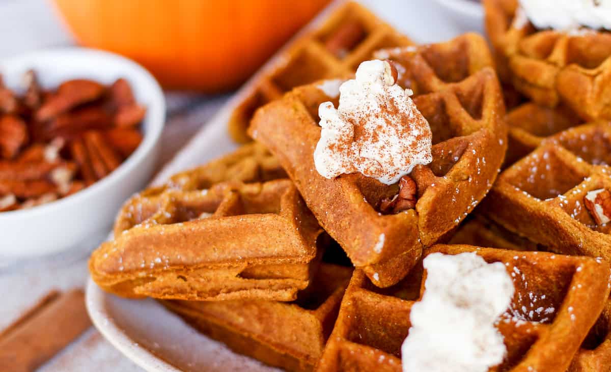 Pumpkin waffles piled on a plate with whip cream on top and a sprinkle of cinnamon.
