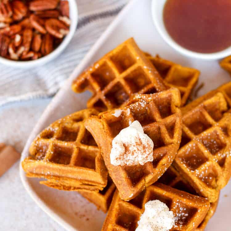 Pumpkin waffles on a white serving platter with small bowls of syrup and pecans on the side.