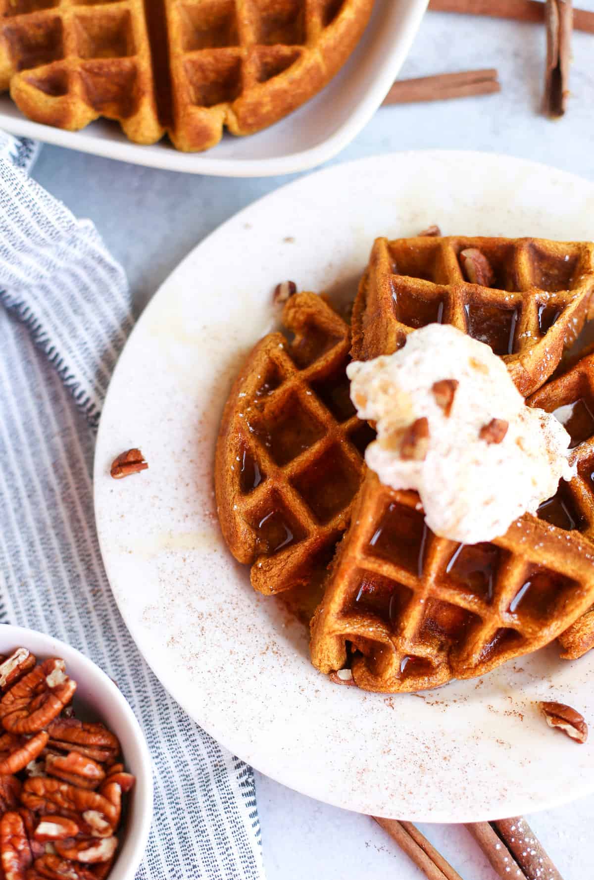 Whole wheat pumpkin waffle on a plate with whipped cream, pecan pieces, and a sprinkle of cinnamon over all of it.