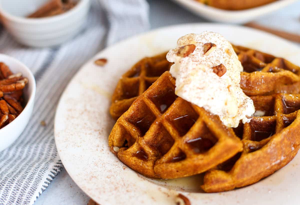 Whole wheat pumpkin waffles on a plate with whipped cream, pecans and cinnamon sprinkled over all of it.