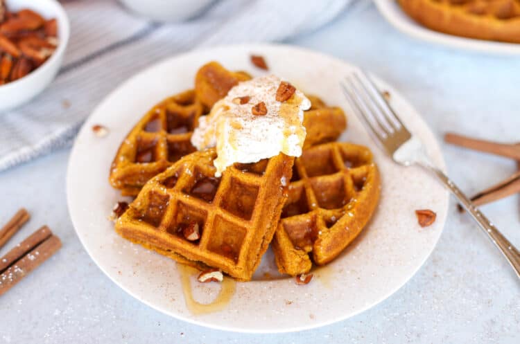 A pumpkin waffle broken in quarters on a plate with syrup, pecan pieces, and a dollop of whip cream on top.
