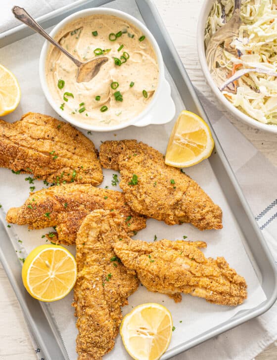 Fried catfish on parchment paper with remoulade sauce and lemon wedges next to them.
