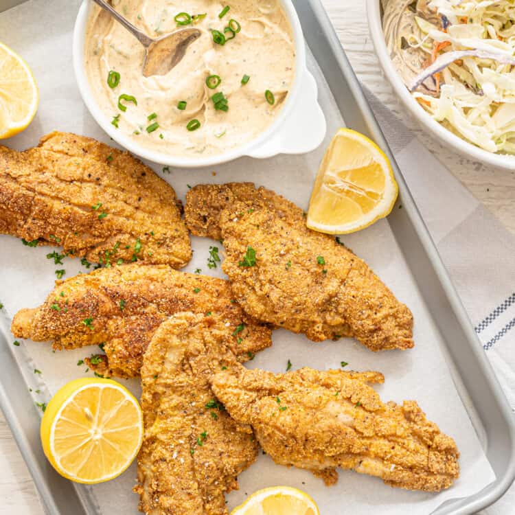 Fried catfish on parchment paper with remoulade sauce and lemon wedges next to them.