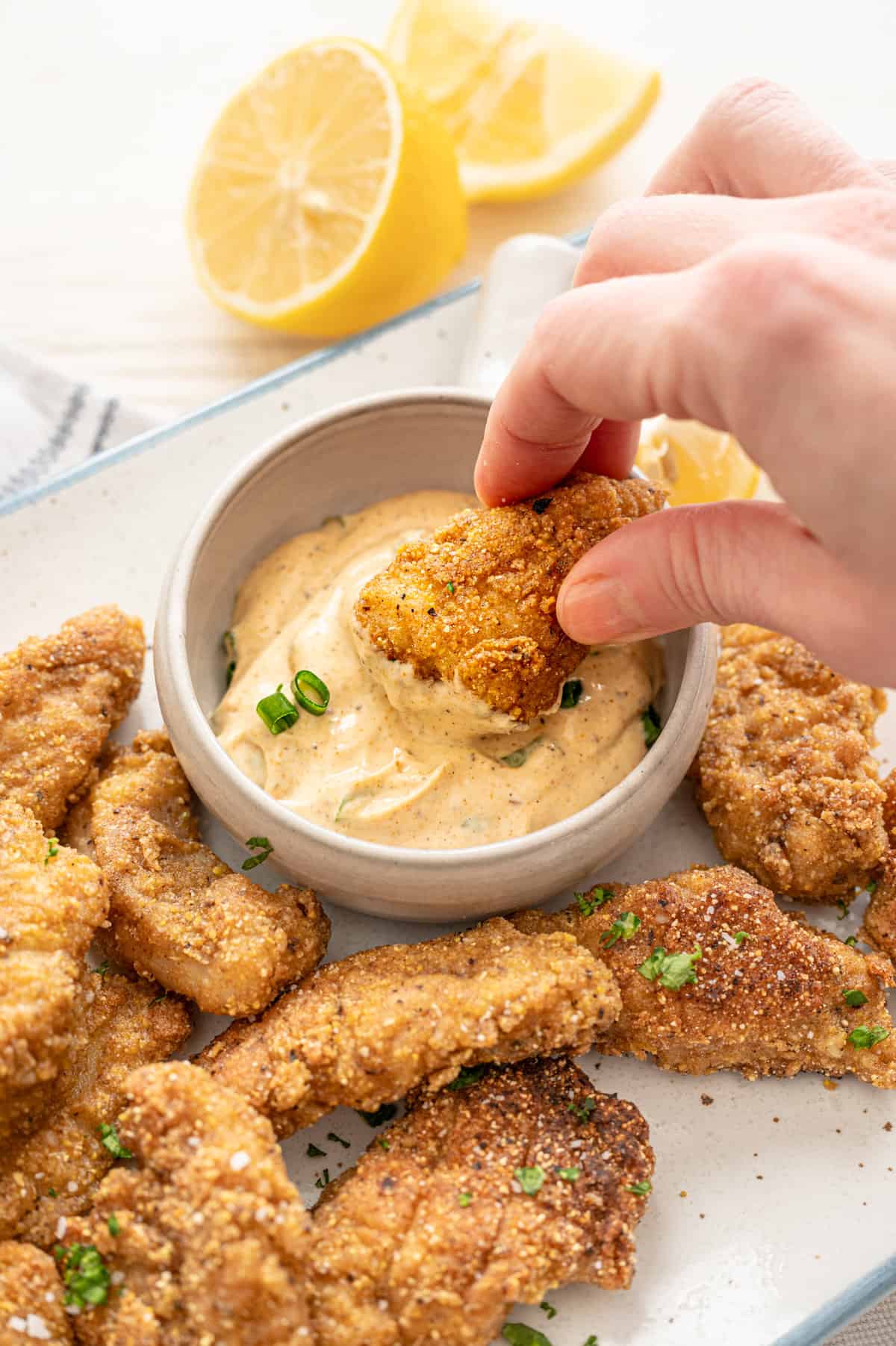 A catfish nugget being dipped in a bowl of Cajun remoulade sauce with other catfish nuggets on a plate.