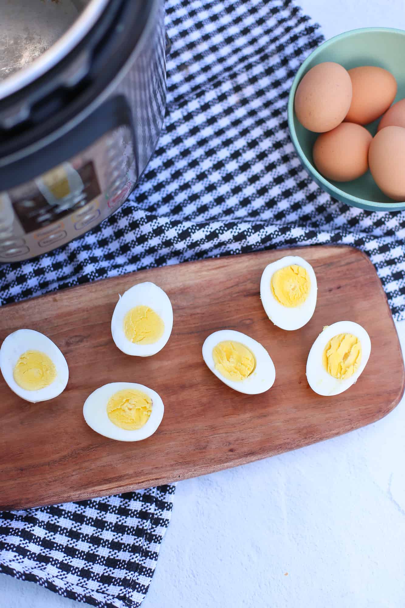 Hard-boiled eggs sliced in half lined up on a wooden tray with an Instant Pot and a bowl of unpeeled eggs in the background.