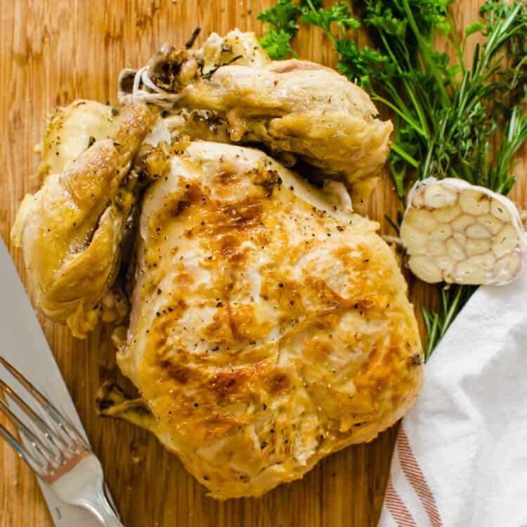 A whole, cooked chicken on a wooden cutting board with a knife and fork nearby.