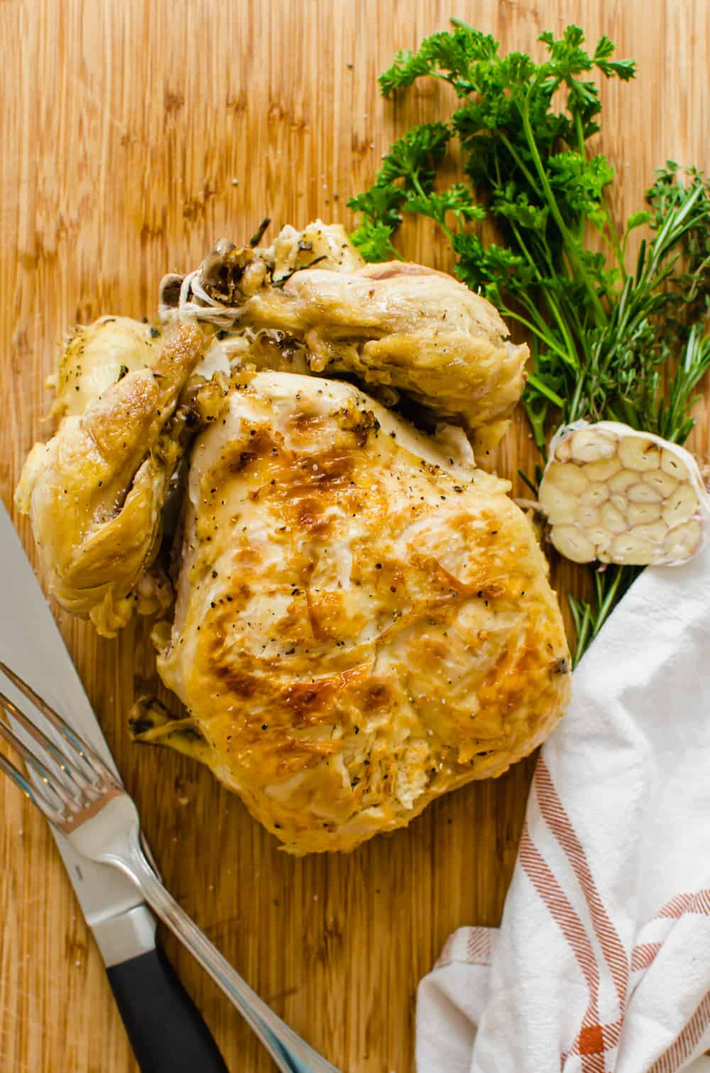 A whole, cooked chicken on a wooden cutting board with a knife and fork nearby.