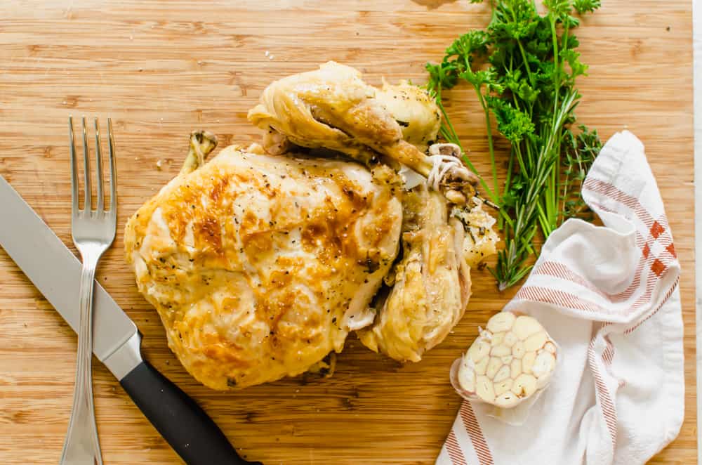 Cooked whole chicken on a wooden carving board with a fork and knife, fresh parsley sprigs and a garlic head cut in half.