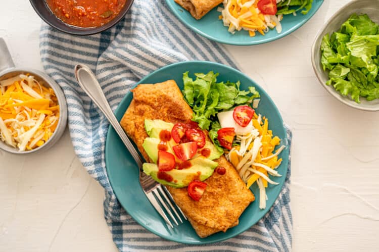 Chicken chimichanga served on a teal plate with chopped tomatoes and avocado on top.