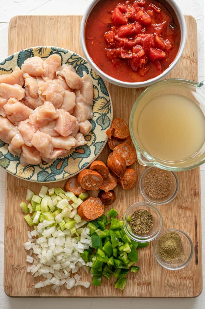 ingredients for jambalaya on wooden cutting board including onion, pepper, seasoning, sausage, chicken, broth, and tomatoes
