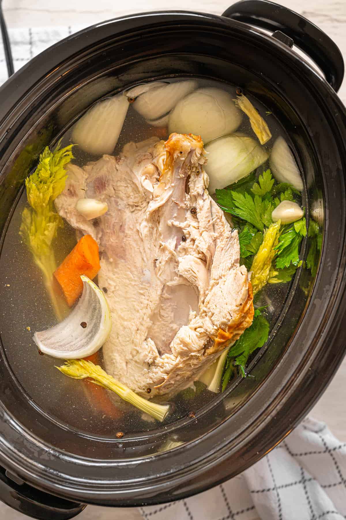 Slow cooker with turkey breast carcass, onions, carrots, celery, garlic cloves, peppercorns, and water to make turkey broth.