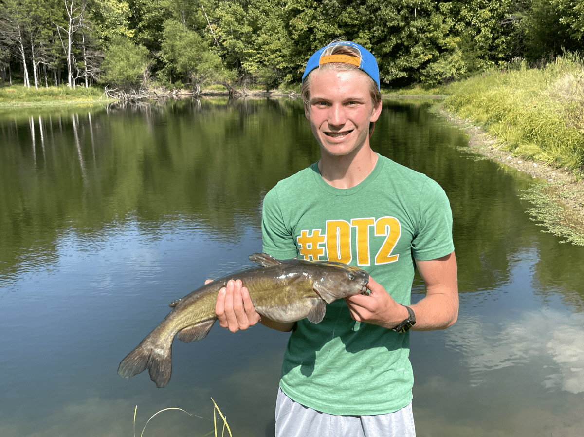 Teenager boy holding a catfish by a pond.