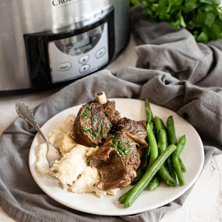Beef short ribs on plate with mashed potatoes and green beans in front of a slow cooker.